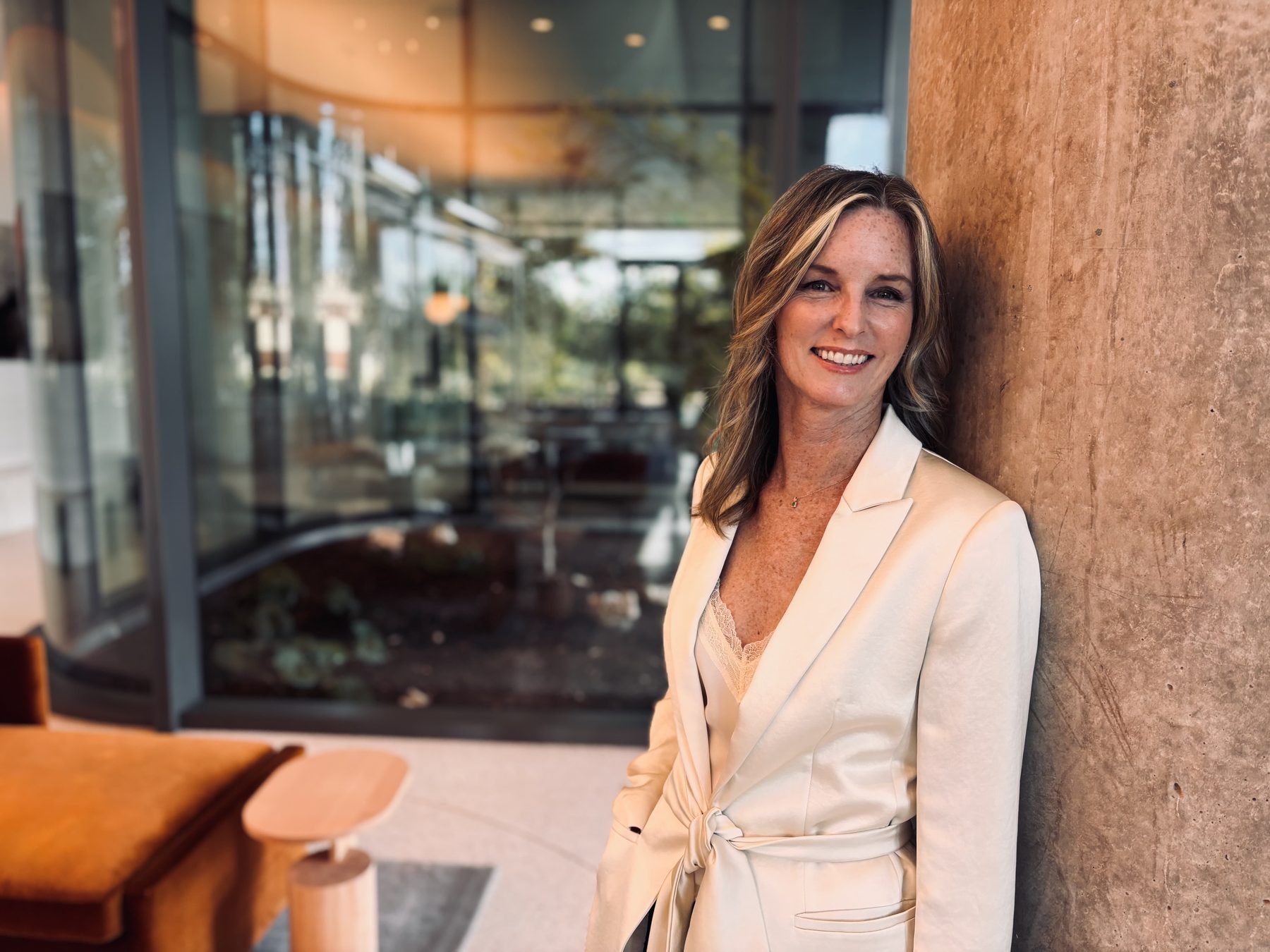 Heather MacLaren smiling, leaning against a warm stone wall in a cream blazer
